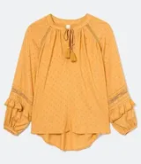 Blusa de manga larga color mostaza con textura de puntos en relieve (dobby). Presenta cuello redondo con abertura en V y cordón ajustable con borla (tassel) en el escote. Las mangas tienen detalles de entretejido calado (entremedios) y volados con terminación fruncida en los puños.