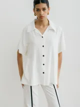 Camisa blanca de corte amplio con cuello clásico y mangas cortas. Cierre frontal con botones negros.