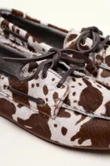 Mocasín tipo boat shoe con estampado de vaca en tonos marrón y blanco, confeccionado en cuero. Cuenta con cordones de cuero marrón oscuro y suela de goma.