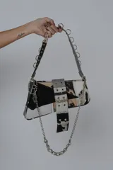 Cartera bandolera de cuero sintético color negro, con diseño patchwork y detalles de tachas y hebillas metálicas. Tiene asa de cadena plateada y cierre con solapa y broche.