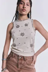 Musculosa color crudo de punto canalé, con cuello redondo y sisas ribeteadas con strass. Presenta apliques de strass con forma de flor y tachas plateadas dispersas por toda la prenda.