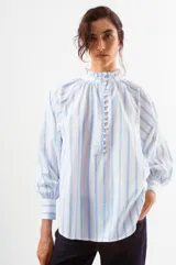 Blusa blanca con finas rayas verticales azules, cuello alto con volado y cierre frontal de botones. Mangas largas abullonadas con puños ajustados.