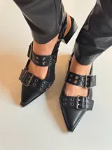 Zapato negro de cuero sintético, con punta fina, escote asimétrico y dos tiras con hebillas metálicas.