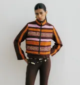 Chaqueta corta con cuello alto y cierre frontal de cremallera. Presenta un diseño a rayas horizontales en tonos marrón oscuro, naranja y rosado. Incluye dos franjas horizontales bordadas con lentejuelas doradas y rosadas en el torso. Las mangas son largas y de color marrón oscuro con una franja naranja vertical.