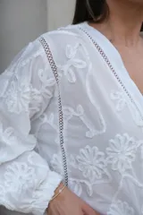 Camisa blanca con bordado de flores, cuello en V, cierre frontal con botones, mangas largas con puños elásticos y detalles de encaje.