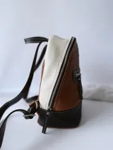 Mochila de cuero color tostado, con base y espalda en negro y tapa superior en blanco. Tiene un bolsillo frontal con cierre y un pequeño aplique de cuero con el logo de la marca.