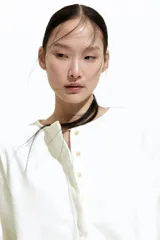 Blusa blanca de corte holgado en sarga de algodón con costuras visibles adelante y atrás. Modelo con cuello redondo, hombros caídos, tapeta con botones ocultos y mangas largas con puños abotonados. Bajo levemente redondeado con abertura pronunciada en los laterales. Espalda algo más larga.