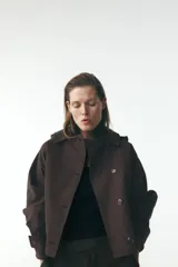 Trench corto color negro, con cuello solapa y capucha desmontable mediante botones. Manga larga tipo murciélago acabada en trabilla ajustable. Bolsillos laterales. Forro interior combinado. Cierre frontal con botones. Tejido repelente al agua.