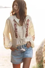 Blusa color crema de cuello redondo y manga larga con elástico en los puños. Presenta bordados florales en el frente y mangas, puntilla a tono y cintura ajustable con cordón y borlas. Cierre frontal con botones.