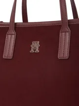Cartera tipo tote bag de color borgoña, con acabado liso y detalles en cuero sintético del mismo tono. Presenta dos asas superiores y una correa ajustable extraíble. Incluye un colgante decorativo con el escudo de Hilfiger bordado y un dije metálico con el monograma TH.