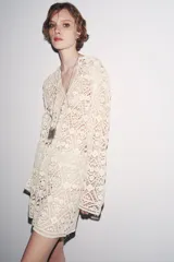 Vestido corto color crudo confeccionado en tejido tipo crochet con diseño geométrico floral. Tiene cuello solapa, abertura con botón en la parte superior y mangas largas acampanadas.