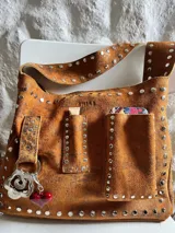 Cartera bandolera marrón de cuero con remaches metálicos plateados. Tiene correa ancha regulable, bolsillo externo y argolla para colgar accesorios.