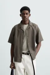 Camisa color marrón oscuro de corte relaxed fit, confeccionada en tejido de lino. Tiene cuello bowling, manga corta, bajo con aberturas laterales y cierre frontal de botonadura.
