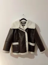Campera marrón de cuero sintético con interior de piel sintética color crema, cuello solapa con hebilla y cierre cruzado con botones a presión.