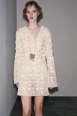Vestido corto color crudo confeccionado en tejido tipo crochet con diseño geométrico floral. Tiene cuello solapa, abertura con botón en la parte superior y mangas largas acampanadas.