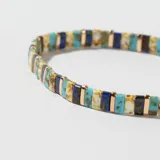 Pulsera elástica con cuentas rectangulares de colores azul, celeste, marrón y blanco, separadas por pequeñas piezas metálicas doradas.