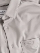 Camisa de manga larga color beige, corte slim, con cuello clásico, cierre frontal con botones, bolsillo de parche en el pecho y logo de Calvin Klein en el bolsillo.