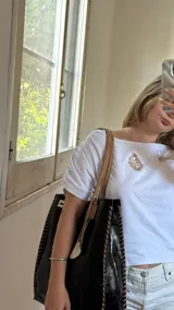 Bolso tote negro de cuero sintético con textura de cocodrilo. Presenta costuras decorativas en los laterales y un dije colgante en forma de cruz plateada con detalles de perlas.