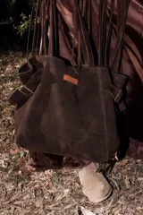 Bolso tote de gran tamaño, color marrón oscuro, confeccionado en material gamuzado. Presenta tiras de hombro largas con flecos colgantes, detalles de hebillas metálicas en los laterales y un parche de cuero marrón claro cosido en el frente.
