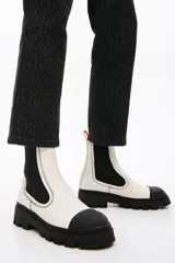 Botines Chelsea de cuero sintético blanco con puntera y suela track de goma negra. Elásticos laterales negros y tirador trasero naranja.