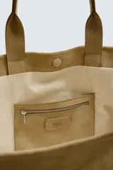 Bolso tipo shopper color beige confeccionado en piel con acabado serraje. Cuenta con doble asa de mano y compartimento principal con cierre magnético. El interior tiene un bolsillo con cierre de cremallera.