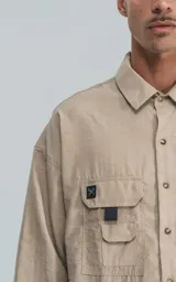 Camisa beige de manga larga, estilo técnico con múltiples bolsillos delanteros y un bolsillo en la parte inferior de la espalda, botones en la cartera y mangas, calce oversize, confeccionada en textil ligero suave al tacto.