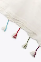Pañuelo rectangular color crudo, confeccionado en lino, con borde color beige y flecos de hilo blanco con detalles de colores.