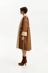 Abrigo largo de diseño envolvente, exterior de cuero gamuzado color marrón y forrería interior de corderito color beige. Presenta solapas anchas de corderito y puños también de corderito.