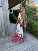 Vestido largo de tejido crochet blanco, sin mangas, con apliques bordados multicolores en el torso y flecos largos teñidos en degradé de blanco a fucsia en el ruedo.