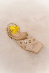 Sandalias color beige con tiras cruzadas adornadas con tachas y ojales metálicos, cierre con pulsera fina y hebilla regulable y taco ancho de 6,5 cm.