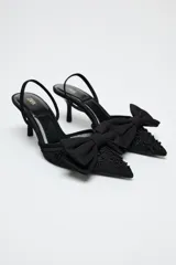 Zapato destalonado negro con tacón fino, capellada de mesh bordado y lazo decorativo en la parte delantera.