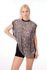 Musculosa corta de jersey con estampado camuflado en tonos verde, marrón y beige. Tiene cuello redondo negro, sisas amplias con ribete negro y estampa lateral con diseño abstracto en negro.