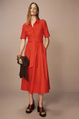 Vestido midi camisero color ladrillo, confeccionado en popelina de algodón. Presenta cuello solapa, manga corta con puños abotonados, bolsillos laterales ocultos y bolsillos de parche con solapa en el pecho. Incluye cinturón lazada en el mismo tejido y cierre frontal con botones ocultos.