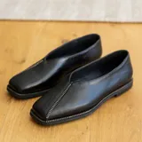 Zapato bajo de cuero negro con punta cuadrada y detalle de cierre en la capellada. Interior forrado en cuero color suela y suela de madera con inclinación de 0,5 cm.
