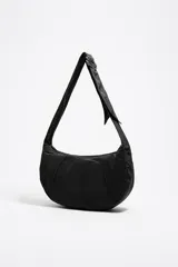 Bolso tipo media luna (hobo bag) de nylon negro, con costuras verticales que le dan estructura y un asa de hombro ajustable con hebilla metálica. Presenta el logo de Bimba y Lola en blanco en la parte inferior central.