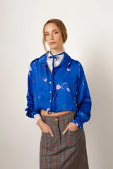 Camisa crop azul con estampado de motivos de fiesta, cuello camisero, mangas largas con puños abotonados y lazos para atar en la cintura.