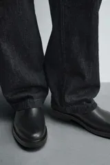 Bota corta estilo Chelsea de cuero liso negro, con elásticos laterales y tirador trasero para calce. Presenta horma redonda y suela con ligero tacón.