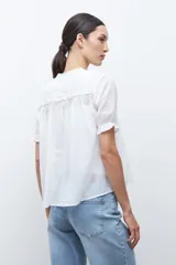 Camisa blanca de manga corta con cuello mao y detalles de bordado en el canesú superior. Presenta frunces en el pecho y la espalda, y un cierre frontal de botones.