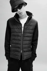 Campera negra híbrida con cuerpo acolchado y mangas de punto. Tiene capucha con cordones ajustables, cierre frontal, bolsillos laterales con botón a presión y puños acanalados.