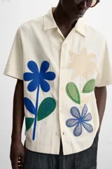 Camisa de manga corta color crudo, con cuello bowling, corte relaxed fit y cierre frontal con botones. Presenta apliques de parches con texturas combinadas a contraste en el delantero y aberturas laterales en el bajo.