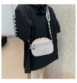 Cartera bandolera pequeña color beige, de cuero sintético con textura granulada. Tiene correa ancha de tela estampada con motivos geométricos y cierre superior con cremallera. Incluye un bolsillo frontal con cremallera y un dije metálico.