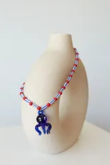 Collar de cuentas de plástico verdes y blancas alternadas, con dije de pulpo rojo de vidrio. El dije de pulpo tiene ojos blancos con pupilas negras.