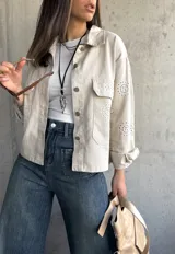 Sobrecamisa color beige con cuello camisero, cierre frontal con botones y bolsillos de parche en el pecho. Presenta detalles de broderie calado en mangas, hombros y bolsillos.