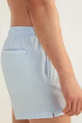 Short de baño celeste con rayas verticales blancas, de sirsaca, con aberturas laterales cortas, elástico en la cintura con cordón ajustable, bolsillos laterales y bolsillo trasero con cierre autoadherente. Suspensor interior de malla.