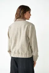 Campera estilo bomber de cuello redondo, color beige jaspeado, con cierre frontal de broches y bolsillos delanteros con ribete marrón.
