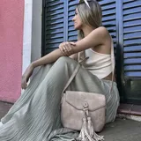 Bandolera color beige con tapa y broche magnético, apertura superior con cierre, detalle de trenzas artesanales y pompones en el frente, correa larga regulable y bolsillos internos.