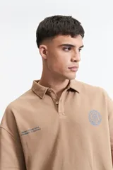 Remera polo color beige con textura acanalada, corte oversize, cuello clásico con botones y logo estampado en el pecho.