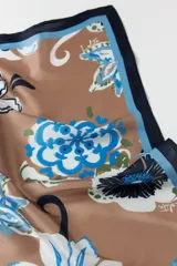 Pañuelo cuadrado de tela color tostado con estampado floral en tonos celeste, azul y blanco. Tiene un borde azul marino con una fina línea celeste.