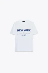 Camiseta blanca de algodón con cuello redondo y manga corta. Estampado frontal con la leyenda "New York" en azul, seguido de la frase "The city that never sleeps EST 1867".