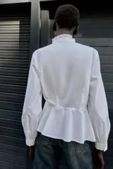 Camisa blanca de popelín con cuello redondo con volante y lazo, manga larga y bajo peplum. Cierre frontal con botones.